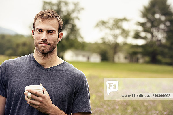 Ein junger Mann mit einer Kaffeetasse in der Hand steht auf einem Feld an einem See.
