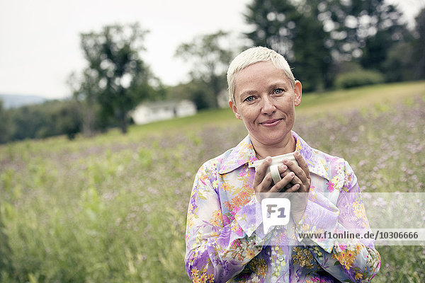 A woman with her hands wrapped around a coffee cup  sitting on a meadow.