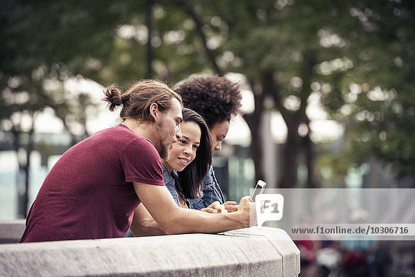 Ein Mann und zwei Frauen  die Smartphones benutzen  stehen in einem Park