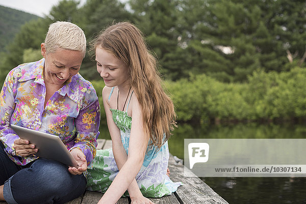 Eine Frau und ein junges Mädchen zusammen auf einem Dock am See mit einem digitalen Tablet.