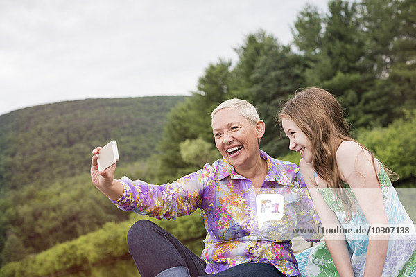 A woman and a child seated on a lake dock taking a selfie with a smart phone.