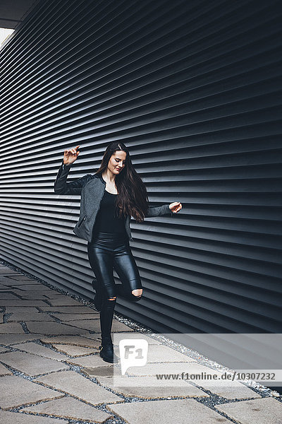 Schwarz gekleidete junge Frau hüpfend auf einem Bein vor schwarzer Fassade