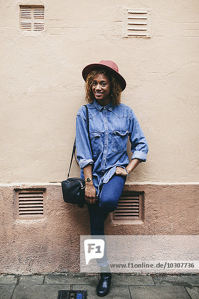 Portrait of smiling young woman wearing hat and denim shirt standing in front of house facade