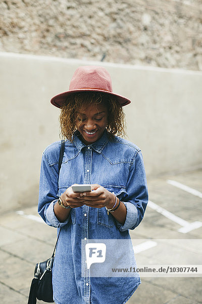 Porträt einer lächelnden jungen Frau mit Hut und Jeanshemd auf dem Smartphone