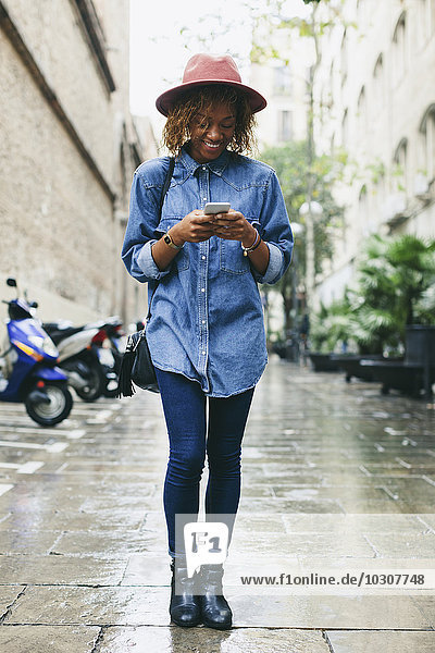 Spanien  Barcelona  Porträt einer jungen Frau mit Hut und Jeanshemd auf ihrem Smartphone