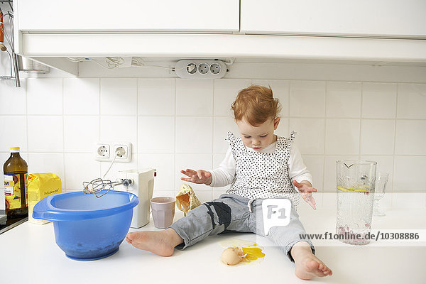 Kleines Mädchen sitzt auf dem Küchentisch und schaut auf gebrochenes Ei.