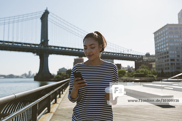 USA  New York City  portrait of smiling young woman with coffee to go looking at her smartphone