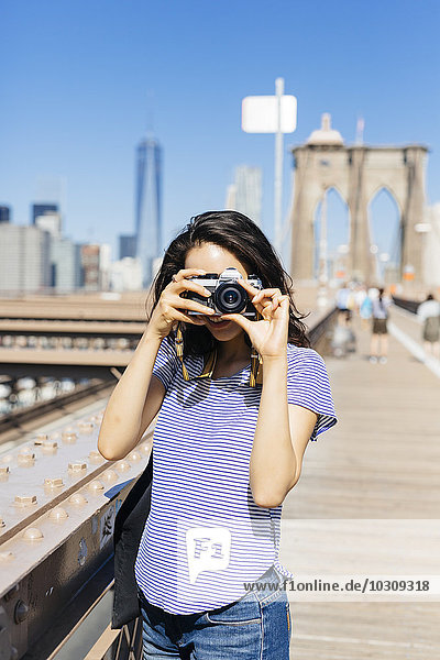 USA  New York City  young woman standing on Brooklyn Bridge taking a photo with camera