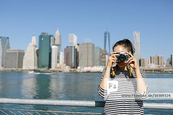 USA  New York City  junge Frau steht vor der Skyline und fotografiert mit der Kamera.