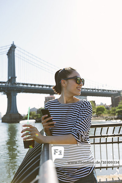 USA  New York City  smiling young woman with soft drink looking at smartphone