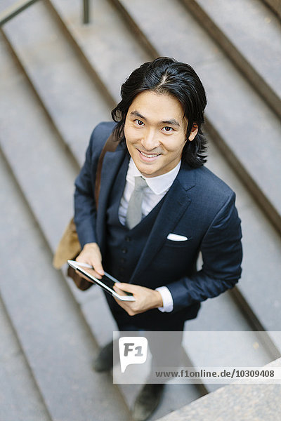 Lächelnder Geschäftsmann steht auf einer Treppe und hält ein digitales Tablett.