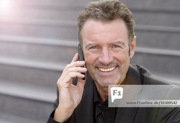 Portrait of smiling businessman sitting on steps telephoning with smartphone