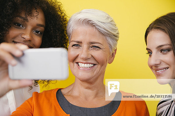 Three women taking a selfie with smartphone in front of a yellow wall