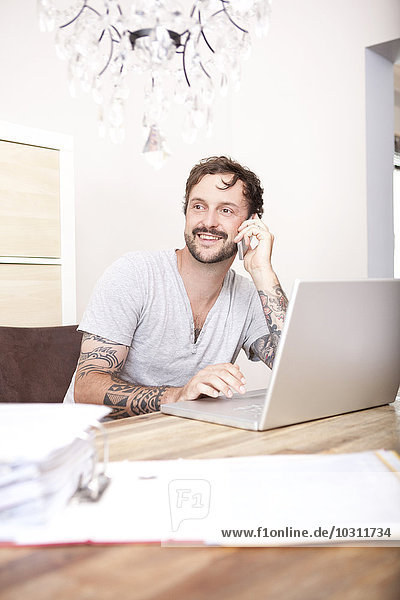 Smiling man sitting at wooden table with laptop and folder telephoning with smartphone