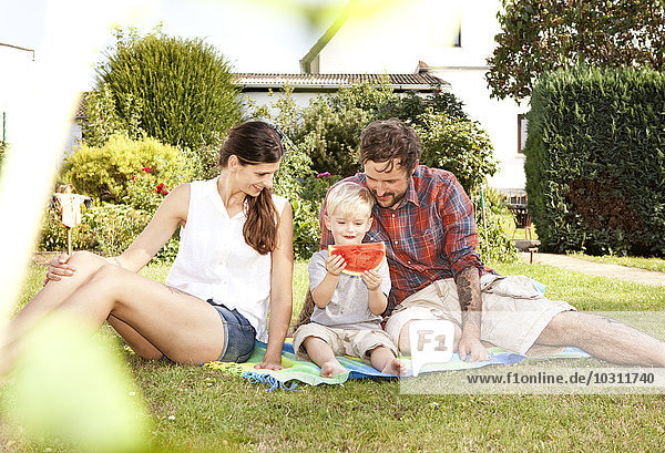 Die Eltern sitzen mit ihrem kleinen Sohn auf einer Decke im Garten.