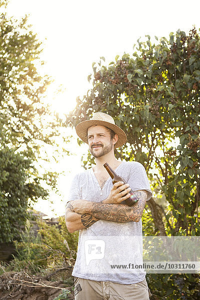 Porträt eines Mannes mit Tätowierungen auf den Armen  der im Garten steht und eine Flasche Bier hält.