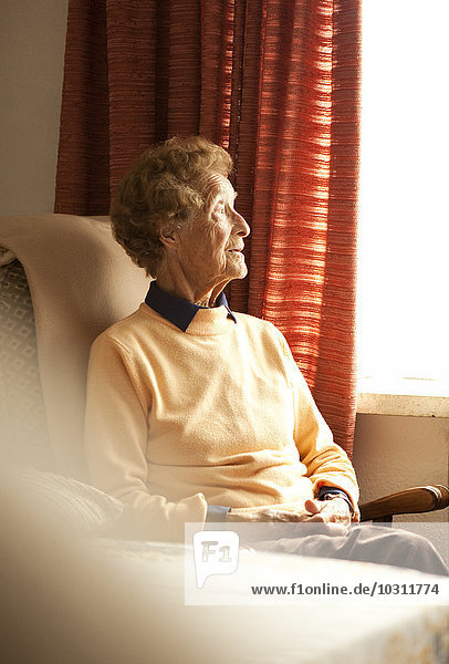 Porträt einer alten Frau  die auf einem Sessel in ihrem Wohnzimmer sitzt und durchs Fenster schaut.