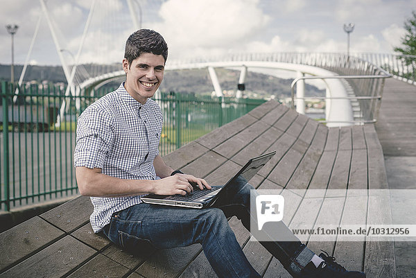 Spain  Ferrol  portrait of smiling young man sitting on a bench with laptop
