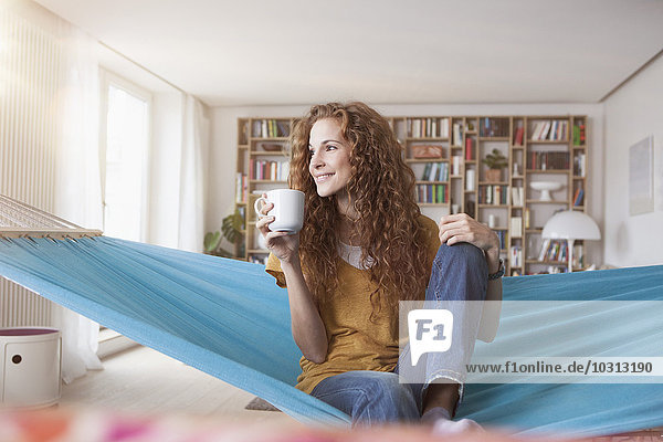 Lächelnde Frau zu Hause sitzend in der Hängematte mit Tasse Kaffee