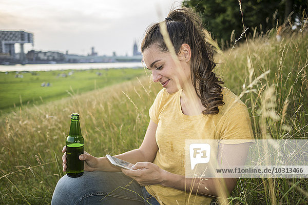 Deutschland  Köln  Frau mit Bierflasche und Smartphone entspannt auf einer Wiese am Rhein