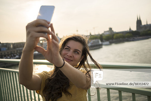 Deutschland  Köln  junge Frau steht auf der Rheinbrücke und nimmt einen Selfie mit ihrem Smartphone.