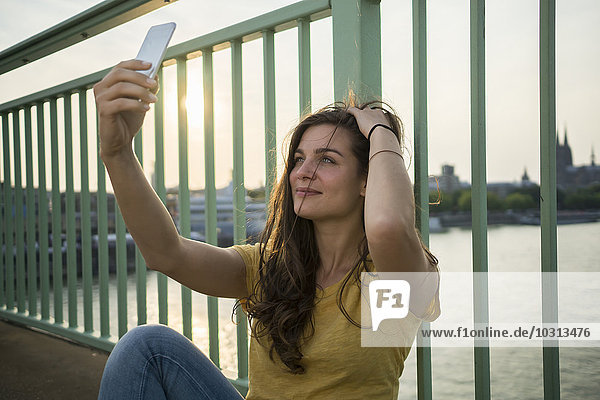 Deutschland  Köln  junge Frau sitzt auf der Rheinbrücke und nimmt einen Selfie mit ihrem Smartphone.