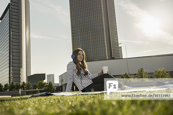 Deutschland  Frankfurt  junge Frau auf einer Wiese sitzend  Musik hören mit Kopfhörer