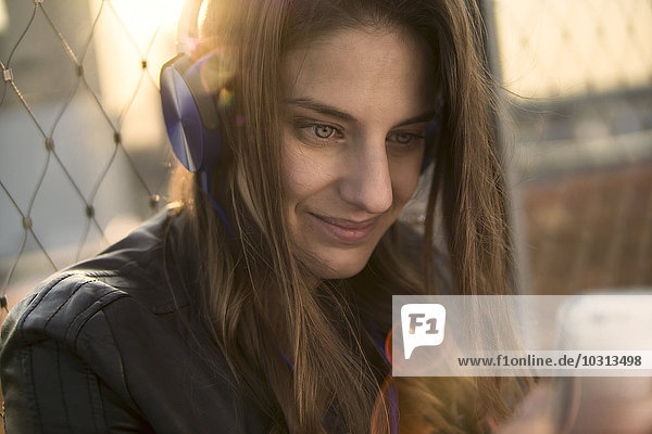 Deutschland  Frankfurt  Porträt einer lächelnden Frau  die Musik mit Kopfhörern hört und auf das Smartphone schaut.