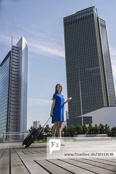 Deutschland  Frankfurt  Geschäftsfrau mit Smartphone und Rollgepäck auf der Dachterrasse stehend
