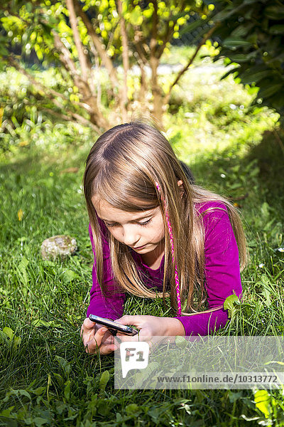 Little girl lying on a meadow in the garden looking at smartphone