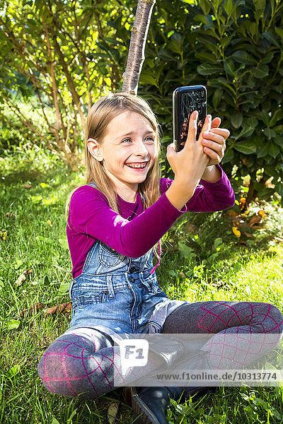 Kleines Mädchen sitzt auf einer Wiese im Garten und nimmt sich ein Selfie mit Smartphone.