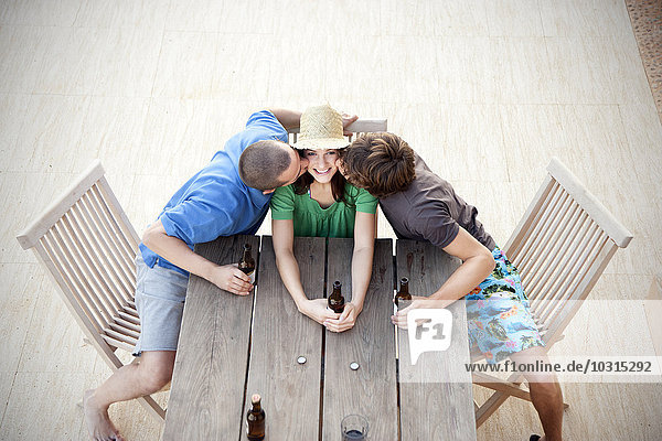 Happy friends sitting at wooden table kissing and drinking beer
