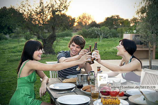 Happy friends sitting at garden table on a barbecue clinking beer bottles