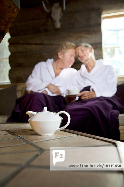 Entspanntes Seniorenpaar auf der Bank im Bademantel sitzend mit Teekanne im Vordergrund