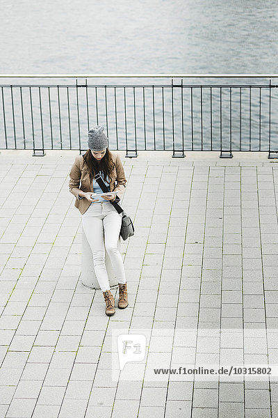 Junge Frau sitzt auf einem Poller an der Promenade und orientiert sich mit einem digitalen Tablett.