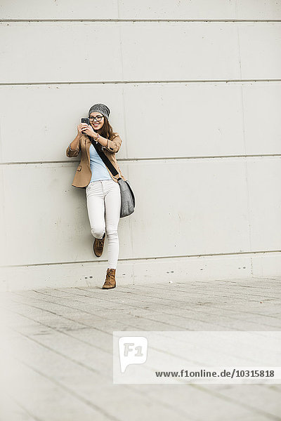 Junge Frau  die sich an eine Wand lehnt und einen Selfie mit ihrem Smartphone nimmt.