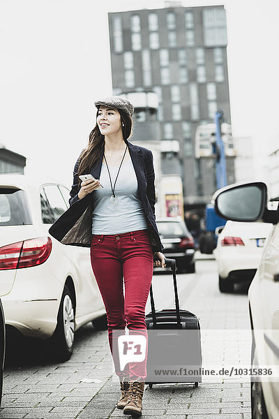 Young smiling woman with smartphone and wheeled luggage