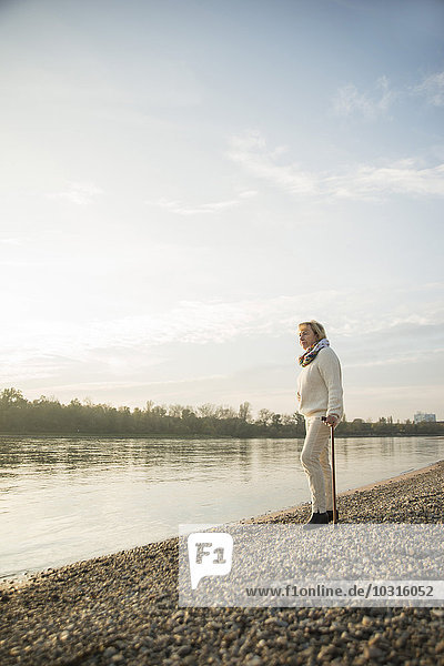 Senior woman with walking stick standing at waterside watching sunset