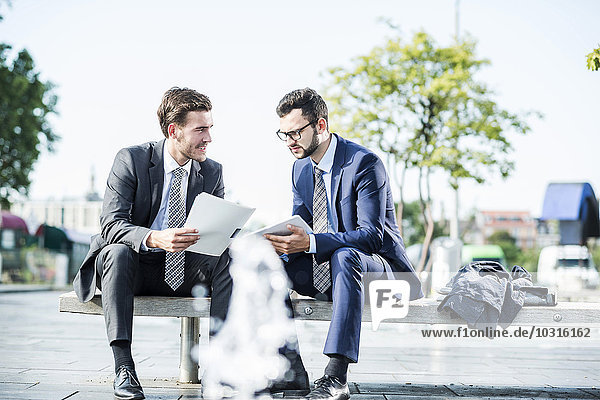 Two young businessmen sitting on bench  discussing files