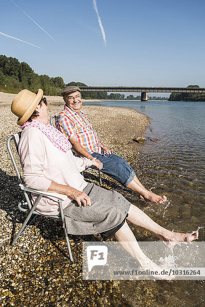 Deutschland  Ludwigshafen  Seniorenpaar sitzt Hand in Hand auf Klappstühlen am Flussufer