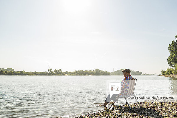 Germany  Ludwigshafen  senior man with beer bottle sitting on folding chair at riverside