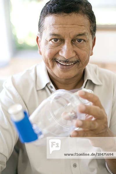 MODEL RELEASED. Asthma spacer use. Man holding an asthma spacer with a blue asthma inhaler. The spacer (plastic chamber) acts as a reservoir  retaining the vapour from the inhaler and allowing the patient to inhale it at their chosen rate. Asthma attacks may be triggered by allergies to dust mites and their faeces. Asthma spacer use