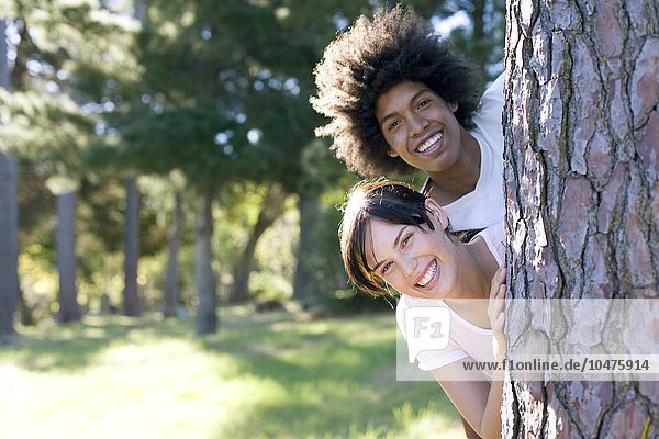MODEL RELEASED. Friendship. Young woman and man looking out from behind a tree trunk. Friendship
