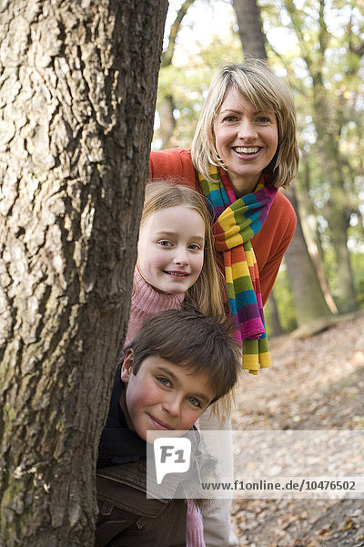 MODEL RELEASED. Playing in a wood. Mother and daughter and son hiding behind a woodland tree in autumn. Mother and children playing in a wood