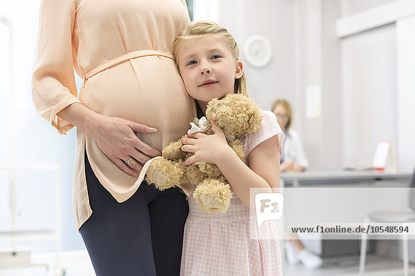 Portrait Mädchen mit Teddybär umarmt schwangere Mutter in Arztpraxis
