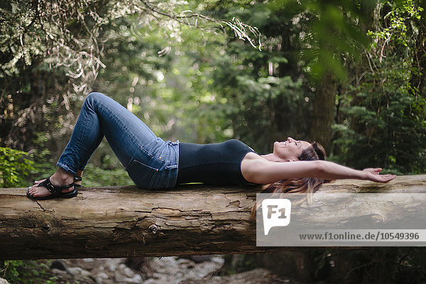 Frau liegt auf dem Rücken auf einem umgefallenen Baumstamm im Wald.
