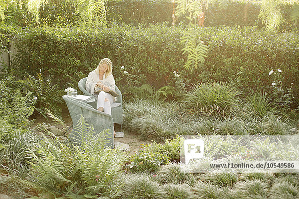 Blonde Frau sitzt in einem Korbstuhl in einem Garten und liest.