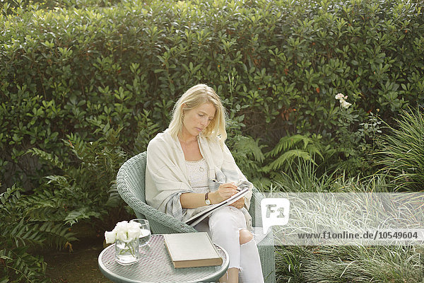 Blonde Frau  die in einem Korbstuhl in einem Garten sitzt und schreibt.
