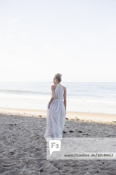 Blond woman wearing a long dress standing on a sandy beach.