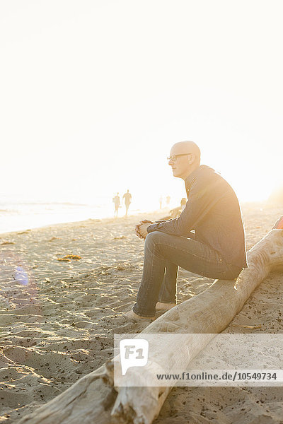 Bald man sitting on a log on a sandy beach.